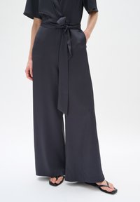Pantaloni larghi grigio scuro con finitura testurizzata, dotati di un dettaglio in vita annodato e tasche laterali, abbinati a ciabatte nere.