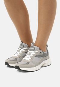 Zapatillas deportivas grises con parte superior de malla y ante, suela blanca gruesa, lengüeta acolchada y acentos en negro contrastante con detalle del logo.