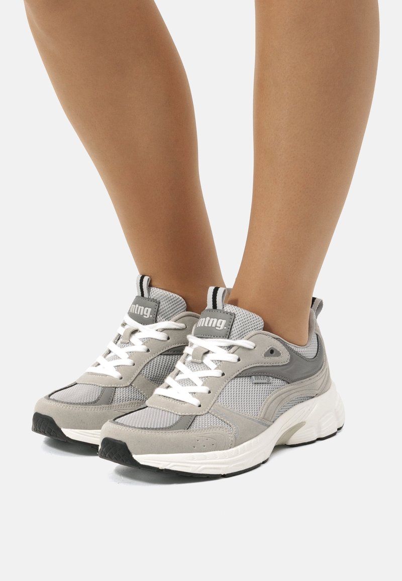 Zapatillas deportivas grises con parte superior de malla y ante, suela blanca gruesa, lengüeta acolchada y acentos en negro contrastante con detalle del logo.