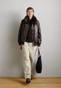 Brun læder bomberjakke med kunstpels krave, beige bukser og sort tote bag. Modellen har kort hår og bærer brune sko.