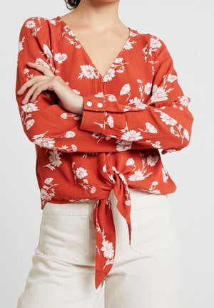 Frau verschränkt die Arme, trägt eine orangefarbene, florale Langarmbluse, die in der Taille gebunden ist, und hochtaillierte weiße Hosen vor einem schlichten Hintergrund.