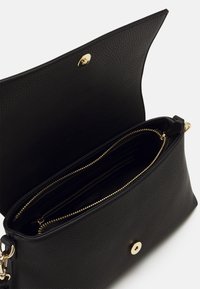 Sac en cuir noir avec surface texturée, équipé d'un compartiment zippé et d'une fermeture à pression dorée. L'intérieur comprend plusieurs poches.