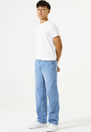 Jonge persoon staand, gekleed in een wit T-shirt, lichtblauwe spijkerbroek en witte sneakers, tegen een effen witte achtergrond.