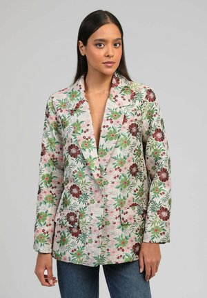 Femme portant un blazer long à motif floral avec des fleurs vertes et bordeaux, associé à un jean bleu, debout devant un fond uni.