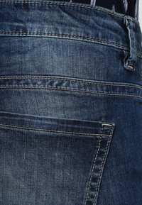 Tmavě modré džíny z denimu s zadní kapsou a viditelnými švy a hladkým, mírně texturovaným povrchem.