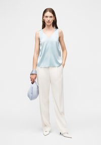 Mujer con top sin mangas de satén azul claro, pantalones anchos blancos, tacones puntiagudos, sosteniendo un bolso y una bufanda a juego en azul claro.