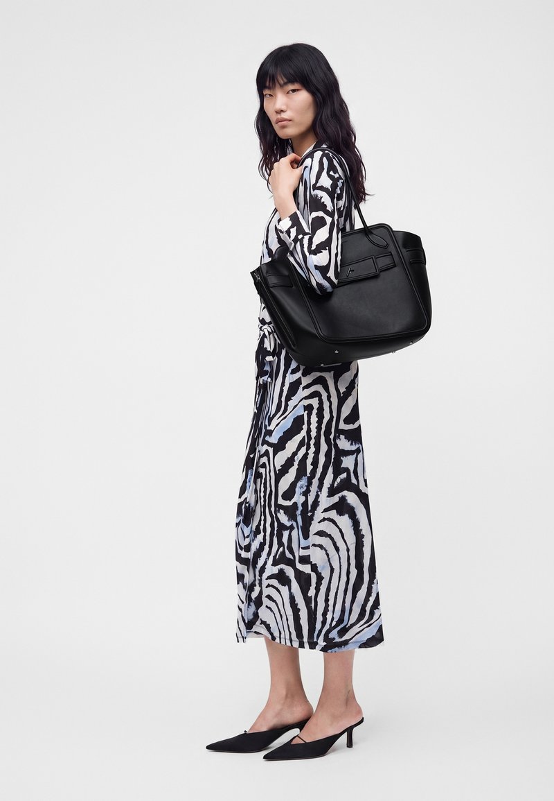 Femme en robe à motif abstrait noir et blanc, tenant un grand sac cabas en cuir noir sur l'épaule, portant des talons pointus noirs.