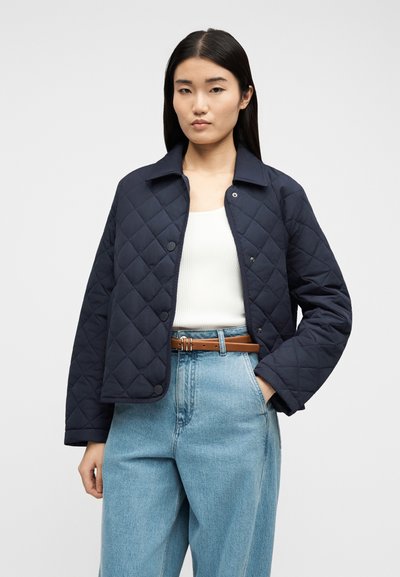 Γυναίκα με μακριά σκουρόχρωμα μαλλιά φοράει μπλε σκούρο μπουφάν με ρίγες πάνω από λευκή μπλούζα, συνδυασμένο με ανοιχτό μπλε τζιν ψηλόμεσο και καφέ ζώνη, με το χέρι στην τσέπη.