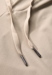 Beige hoodie fabric with textured surface, featuring light beige drawstrings and metal tips. Flat stitching visible on the material.
