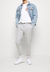 Man i ljusblå denimjacka, vit grafisk t-shirt, ljusgrå randiga byxor, vita strumpor och vita sneakers som står mot en enfärgad bakgrund.