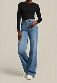 Personne portant un jean bleu taille haute à jambes larges, un haut noir à manches longues et des chaussures blanches, debout avec une main sur la hanche.