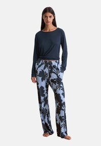 Femme portant un haut à manches longues bleu marine et un pantalon léger bleu clair ample avec de grands motifs floraux noirs, se tenant pieds nus avec les mains dans les poches.