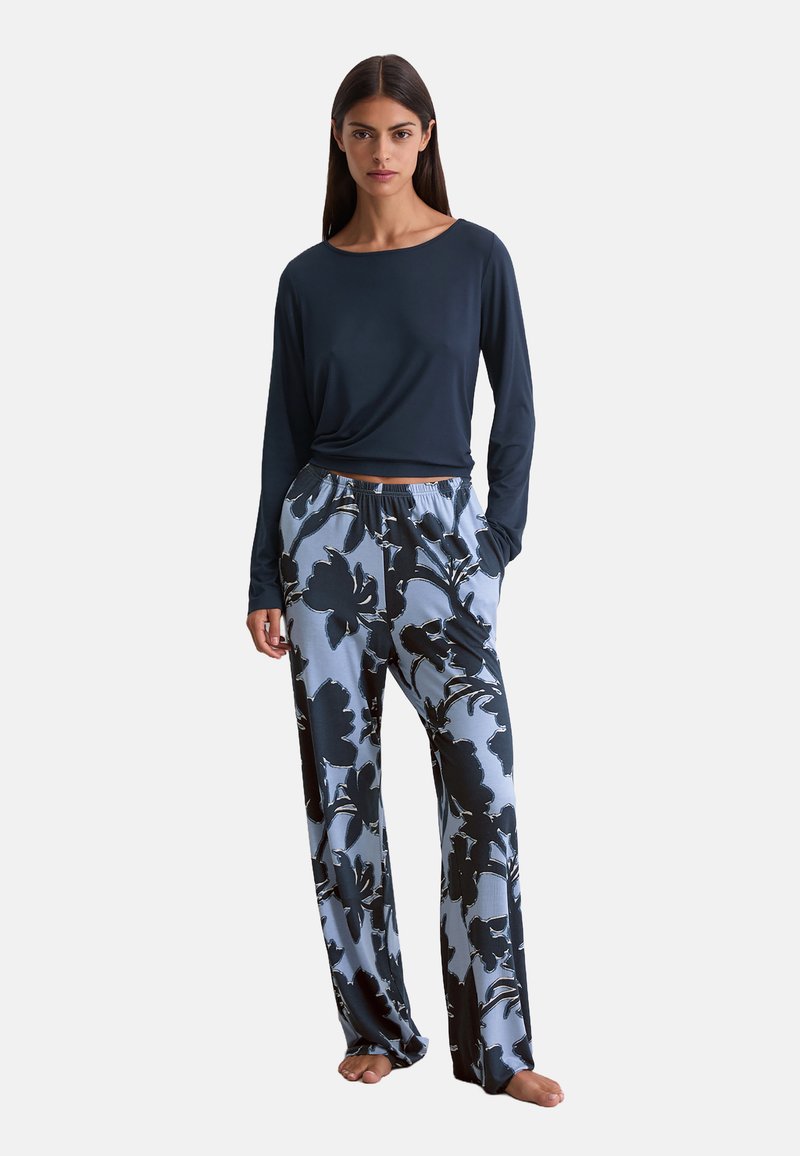 Femme portant un haut à manches longues bleu marine et un pantalon léger bleu clair ample avec de grands motifs floraux noirs, se tenant pieds nus avec les mains dans les poches.