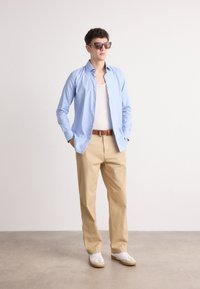 Camisa de mangas longas azul clara, top branco, calças bege, cinto castanho, ténis brancos com detalhes bege e óculos de sol oversized.