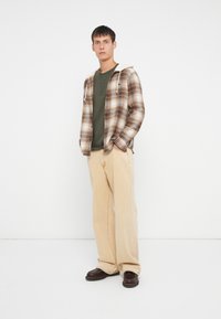 Chemise en flanelle à carreaux marron et crème, pantalon beige doux, henley vert en dessous, et chaussures marron foncé. Le modèle se tient contre un fond blanc.