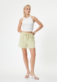 Hvid ribbet tanktop og lysegule shorts med flæset talje og snørelukning. Model iført hvide flip-flops foran en ensfarvet baggrund.