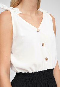 Haut sans manches blanc avec un col en V, présentant des boutons décoratifs et des liens sur les épaules. Le bas a une texture froncée et élastique.