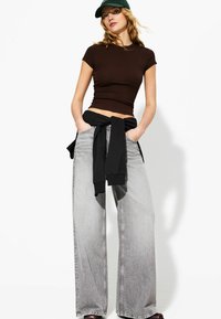 Bruine getailleerde t-shirt, grijze wijdvallende jeans en een zwarte sweatshirt om de taille geknoopt. Accessoires zijn een groene pet.