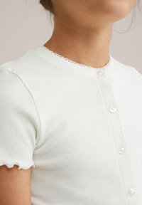 Dijete nosi bijelu rebrastu majicu kratkih rukava s malim gumbima i valovitim rubovima na ovratniku i rukavima, okrenuto bočno.