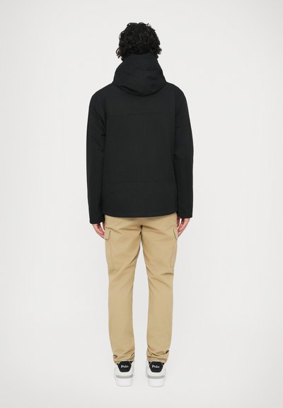 Jachetă neagră cu glugă, cu un finisaj neted, asociată cu pantaloni cargo bej. Adidas albi cu accente negre și branding "Polo" pe călcâi.