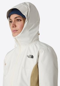 Vit vattentålig jacka med beige accenter, dragkedja fram, hög krage och huva. Har en liten svart logotyp på bröstet.