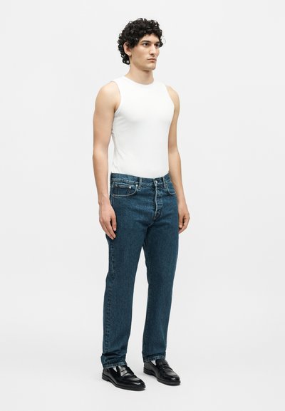 Άνδρας που στέκεται, φοράει λευκό αμάνικο πουκάμισο, μπλε τζιν και μαύρα δερμάτινα παπούτσια, κοιτάζοντας ελαφρώς δεξιά σε απλό φόντο.