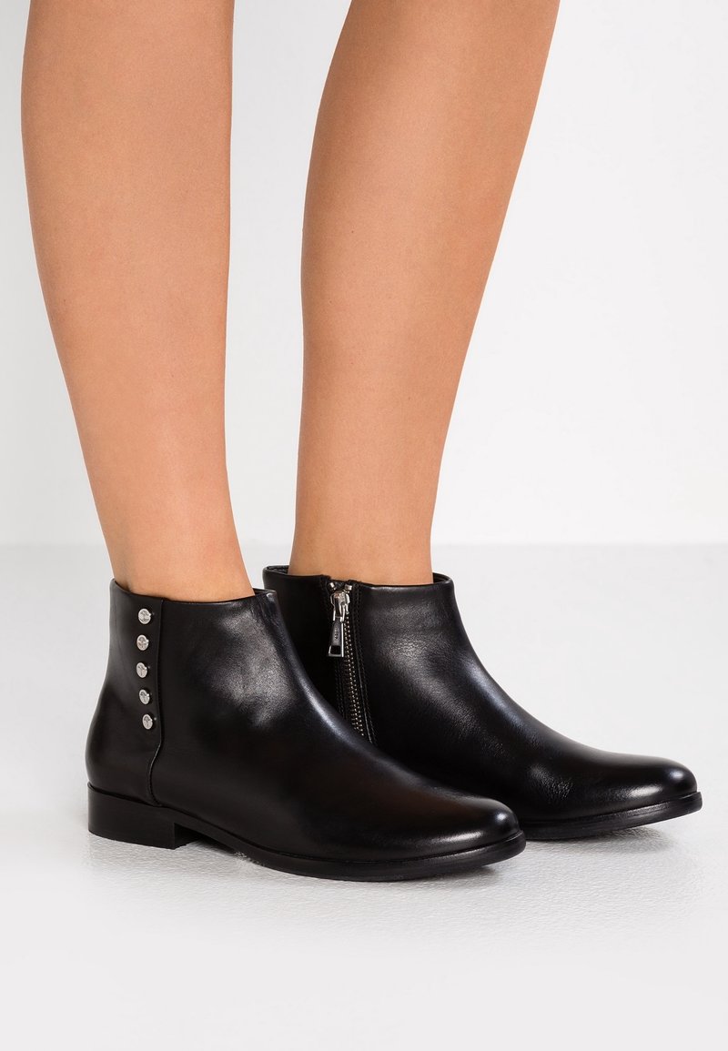 Bottes de cheville en cuir noir avec fermeture éclair latérale et boutons argentés décoratifs portées sur des jambes nues sur fond blanc.
