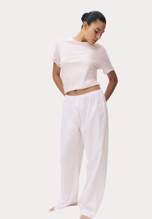 Vrouw met donker haar in een knot, gekleed in een lichtroze top met korte mouwen en losse witte broek, staat op blote voeten tegen een effen achtergrond.