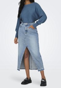 Jupe en jean bleu clair avec une fente à l'avant, taille haute et coupe droite. Associée à un pull bleu côtelé et des chaussures noires.