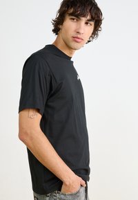 Camiseta de algodón negra con manga corta, cuello redondo y un sutil logo blanco en el pecho. La tela parece suave y ligera.