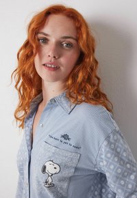 Lys blå skjorte med knapper, der har vertikale striber og blomsterdesign. Forkert lomme inkluderer et broderet Snoopy-design og tekst.