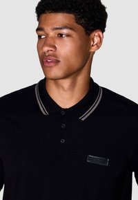 Zwarte poloshirt van een textiel met structuur, met een gestreepte kraag in het grijs, drie knopen placket en een klein logo op de borst.