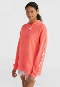 Korale różowy hoodie z kieszenią kangurzą, kapturem ze sznurkiem i białym napisem "Kobiety Fali" wzdłuż rękawa. Miękka tekstura materiału.