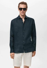 Camisa de botones de manga larga en lino de color azul oscuro con una textura sutil. Combinada con pantalones cortos beige claro. Lleva unas gafas de sol negras.