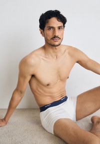 Jeune homme assis aux cheveux foncés portant un boxer blanc avec une ceinture bleu marine, reposant sur un sol moquetté clair contre un mur uni.