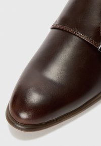 Zapato de piel marrón con un acabado suave, presentado con punta afilada y una correa con una hebilla plateada. Costuras decorativas visibles a lo largo de los bordes.