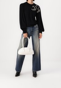 Jersey negro con mangas abullonadas y bordado floral beige, combinado con unos vaqueros de pierna ancha color azul claro y un bolso blanco curvado con doble asa.
