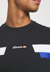 Zbliżenie na osobę w czarnej koszulce z białymi i niebieskimi kształtami geometrycznymi oraz naszywką z logo ellesse na piersi.