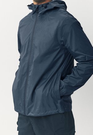 Mężczyzna w ciemnoniebieskiej kurtce przeciwdeszczowej z kapturem, zamkiem błyskawicznym i rękami w bocznych kieszeniach, na jednolitym tle.
