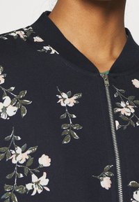 Květinová bomber bunda s navy pozadím, která má růžové a zelené květinové vzory, žebrovaný límec a stříbrný zip.
