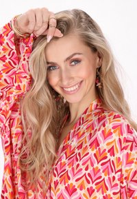 Tela con un patrón floral colorido y motivos de corazón en rosa, naranja y rojo. La modelo tiene el cabello largo y rubio, y lleva puesto joyería de oro.