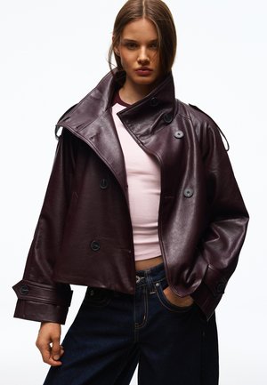 Jeune femme portant une veste en cuir double boutonnage bordeaux foncé sur un haut rose clair et un jean bleu foncé, posant avec une main dans la poche.