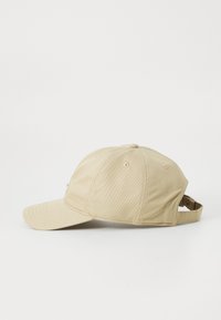 Beige keps i tyg med en strukturerad skärm, med en justerbar rem bak och subtila sömmar. Enkelt design och texturerat material.