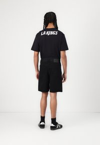 Svart T-skjorte med "LA KINGS" trykk på ryggen, kombinert med svarte denimshorts. Modellen har på seg svarte sokker og sko, sett fra bak.