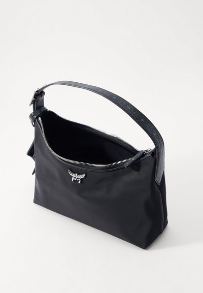 Melnas pleca soma ar platu teksturētu siksnu, daļēji atvērtu rāvējslēdzēju un mazu sudraba emblēmu priekšpusē uz balta fona.