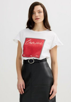 Mujer con pelo largo y castaño, vistiendo una camiseta blanca con estampado rojo y una falda de cuero negra de talle alto con cinturón de hebilla redonda.