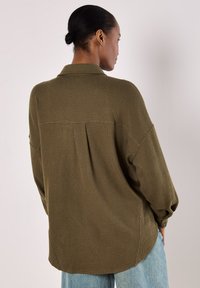 Olive green, textured, long-sleeve shirt featuring a collar, back pleat, and rounded hem. Styled with light blue denim pants.