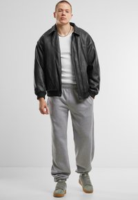 Veste en cuir noir de type bombardier, débardeur blanc, pantalon de survêtement gris et baskets vertes avec une semelle en gomme. Le modèle se tient devant un fond uni.