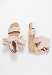 Beżowe sandały na koturnie z materiałowymi sznurkami wokół kostki i pojedynczym paskiem z przodu, jeden pokazany z góry, a drugi z boku.
