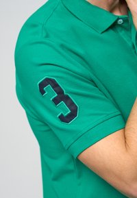 Zelená polo košeľa z textúrovanej bavlny s tmavomodrým vyšitým číslom "3" na krátkom rukáve. Disponuje klasickým designom goliera.